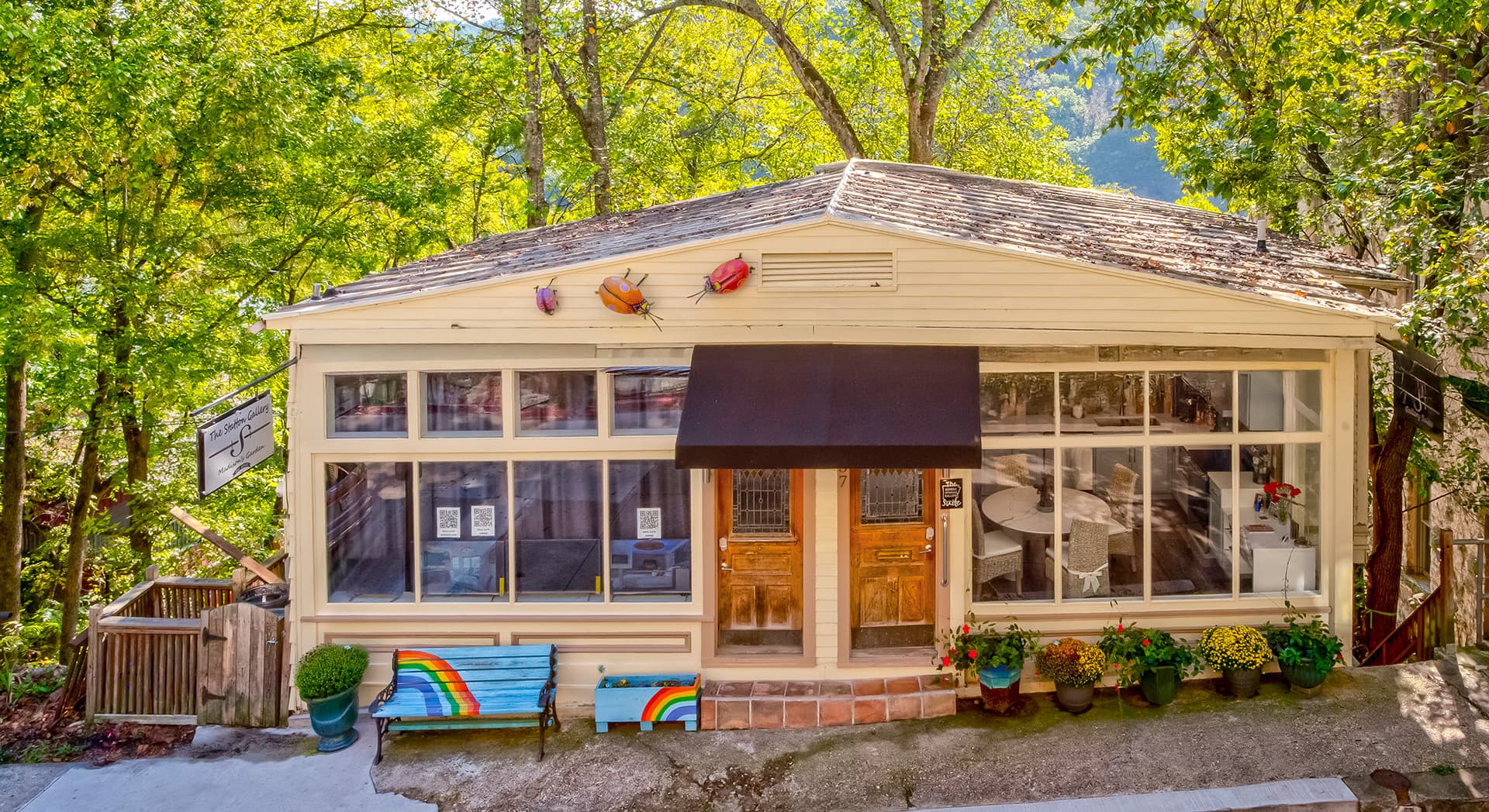 A charming single-story building with large windows, a black awning, and colorful flower pots, surrounded by lush greenery.