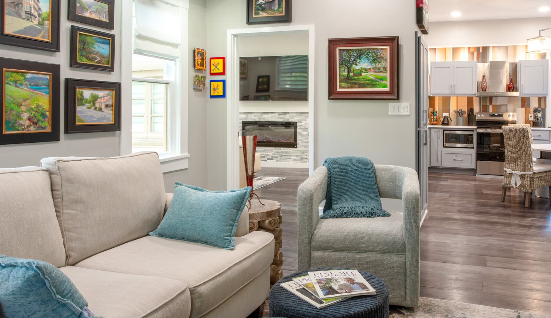 A cozy living room featuring a light sofa, decorative paintings, and a view into a modern kitchen.