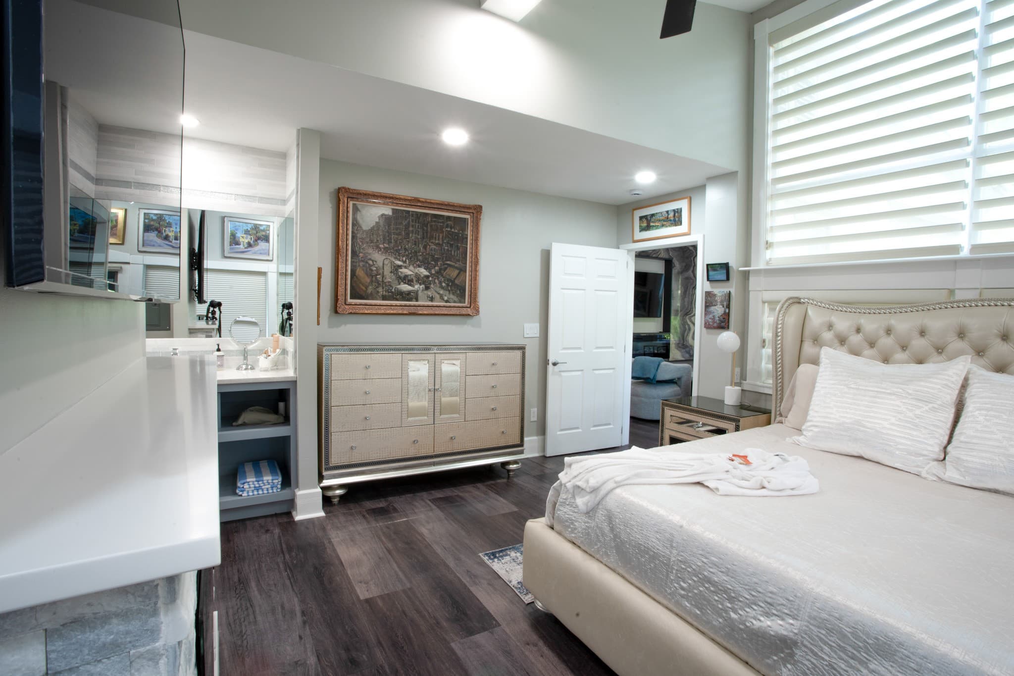 A modern bedroom featuring a large bed, a dresser, and a bathroom vanity, with sunlight streaming through slatted windows.