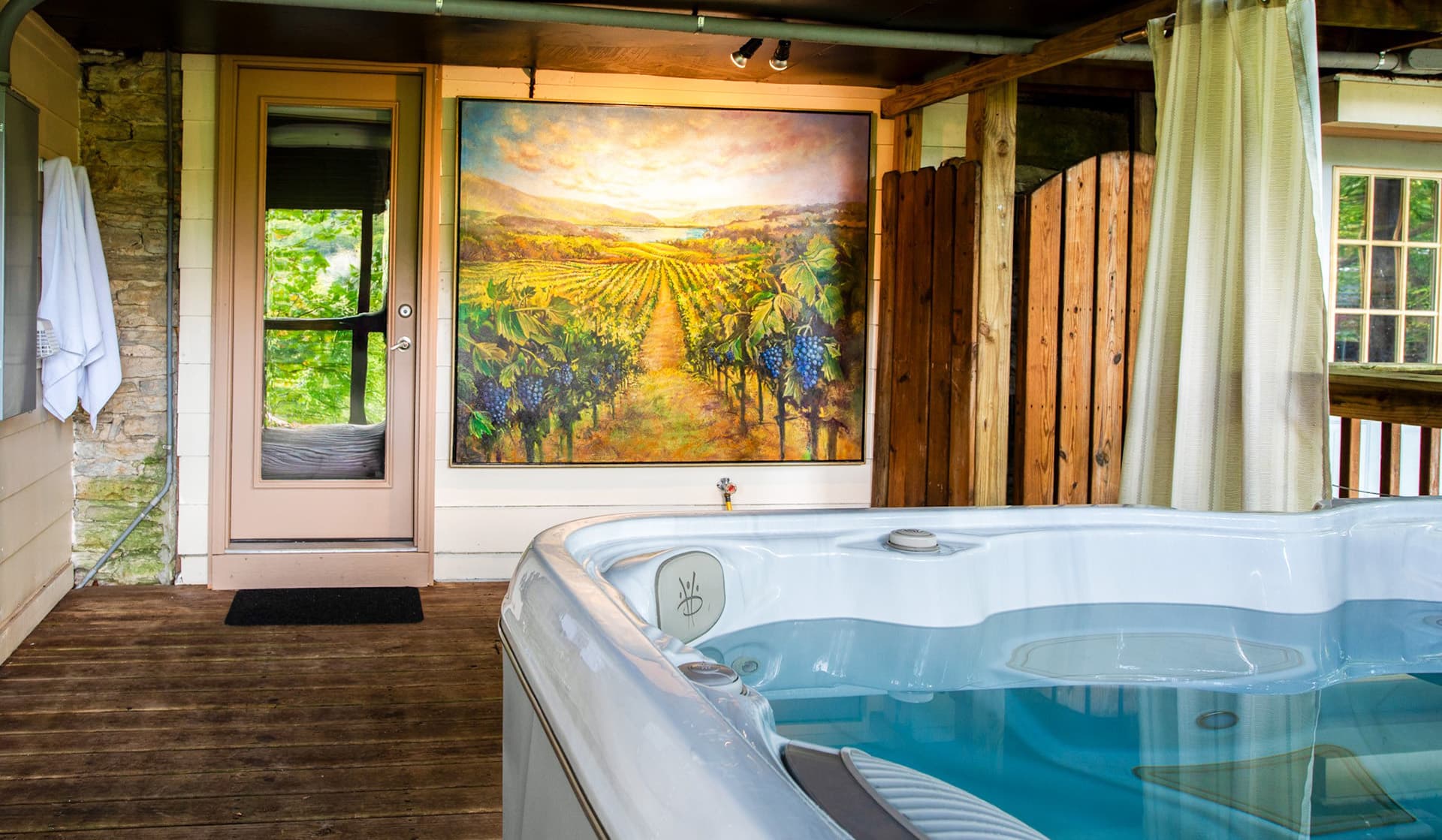 A hot tub on a wooden deck with a vineyard painting and a door leading outdoors.