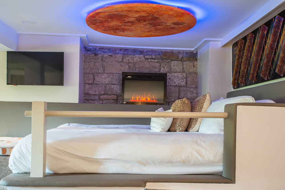 A modern bedroom featuring a large bed, a stone wall with an electric fireplace, and colorful ceiling lighting.