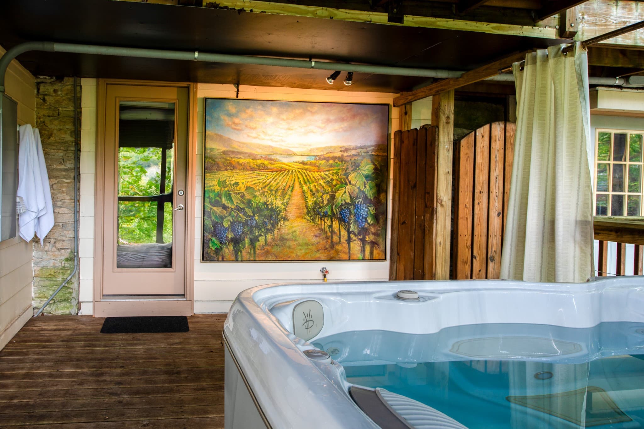 A hot tub on a wooden deck next to a door, with a large vineyard painting on the wall.